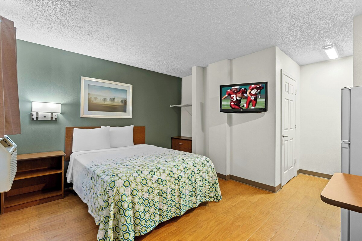 A hotel room with a bed, a TV, a painting, a nightstand, and a wooden floor.