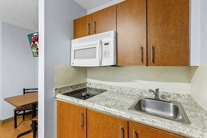A kitchen with wooden cabinets, a white microwave, a granite countertop, and a stainless steel sink. - HomeTowne Studios by Red Roof Orlando South (Orlando)