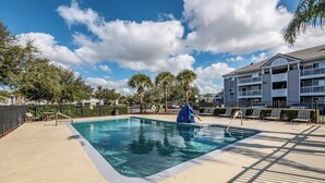 A swimming pool with a deck, lounge chairs, palm trees, and a multi-story building in the background. - HomeTowne Studios by Red Roof Orlando South (Orlando)