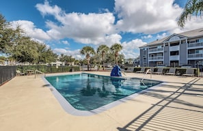 A swimming pool with a deck, lounge chairs, palm trees, and a multi-story building in the background. - HomeTowne Studios by Red Roof Orlando South (Orlando)