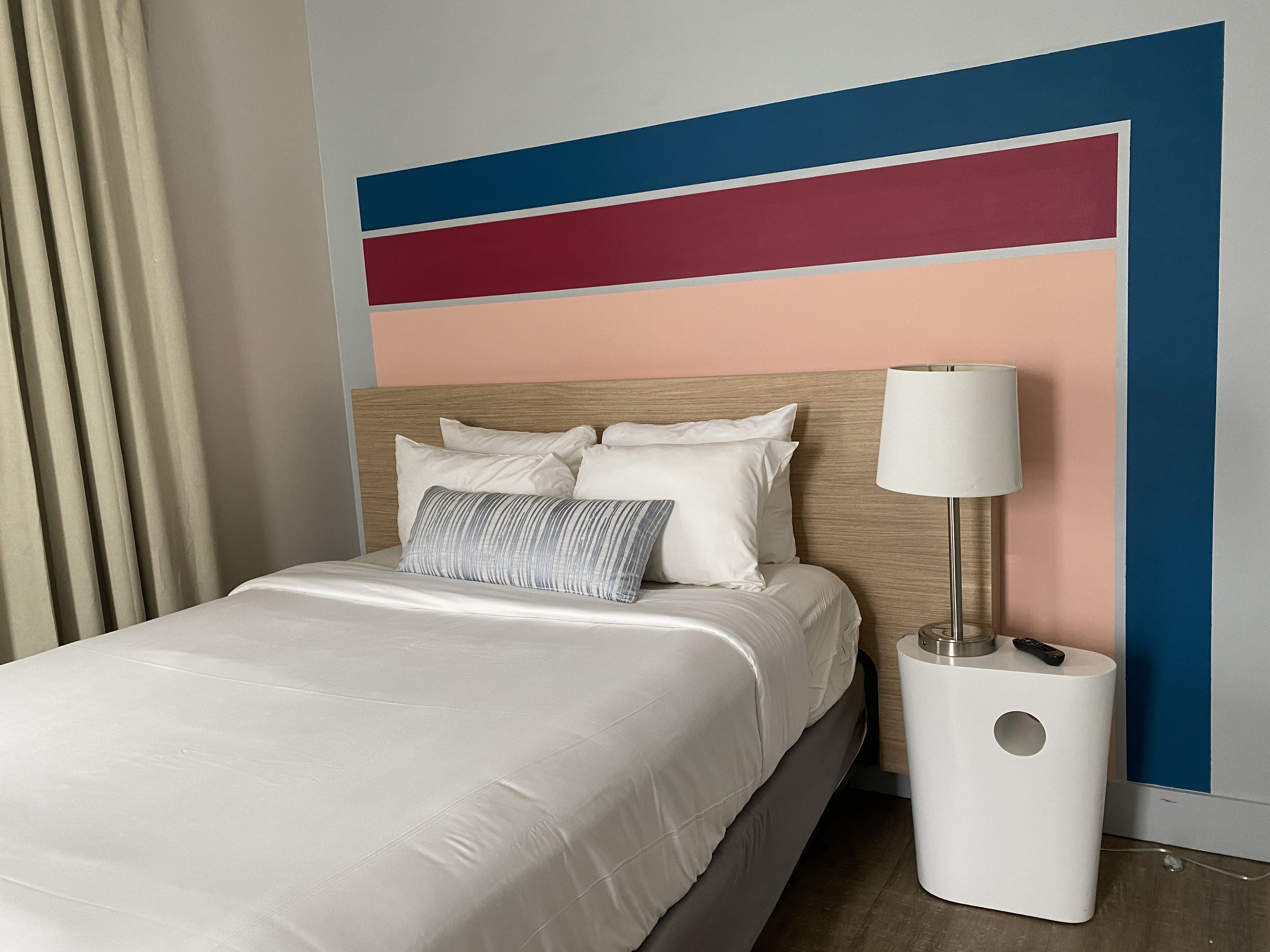 Una cama bien hecha con almohadas, una lámpara de noche y un control remoto sobre una mesita blanca.