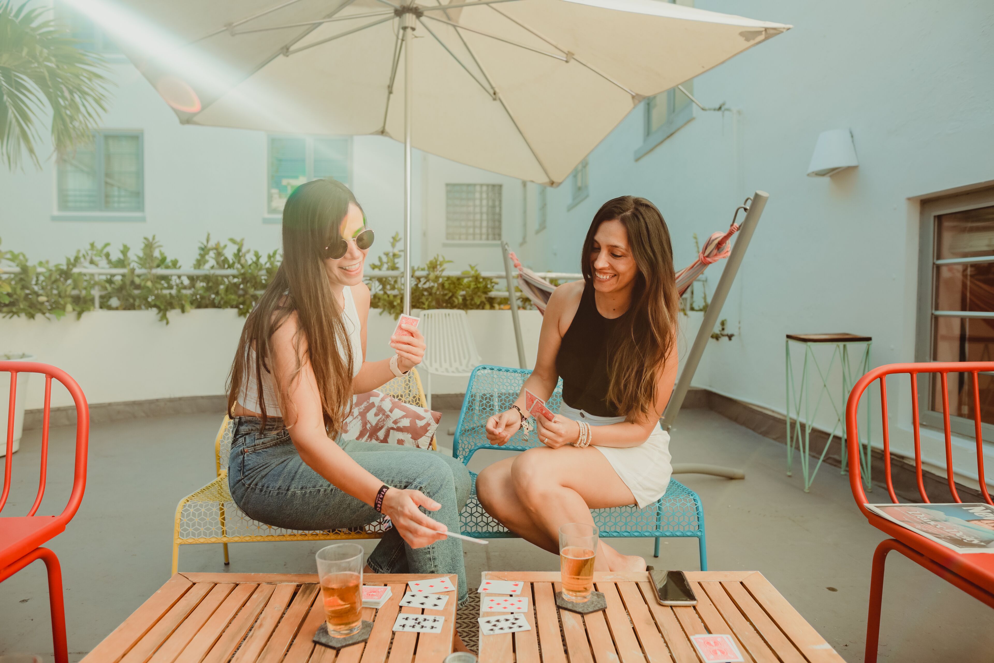 Two people playing cards at an outdoor table with a wooden table, chairs, and an umbrella.