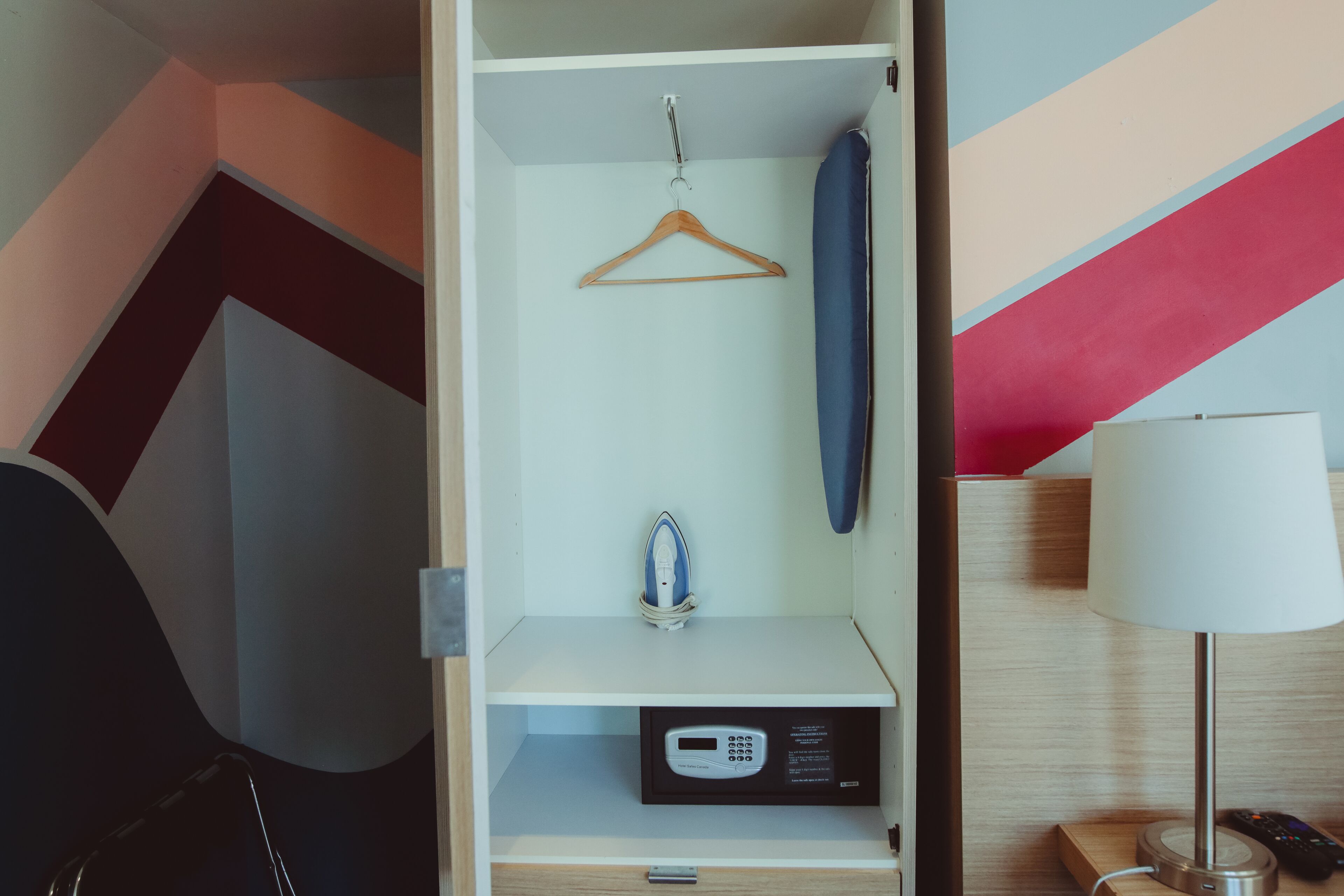 A hotel room with a wardrobe, a hanging coat, an iron, a digital clock, and a lamp.