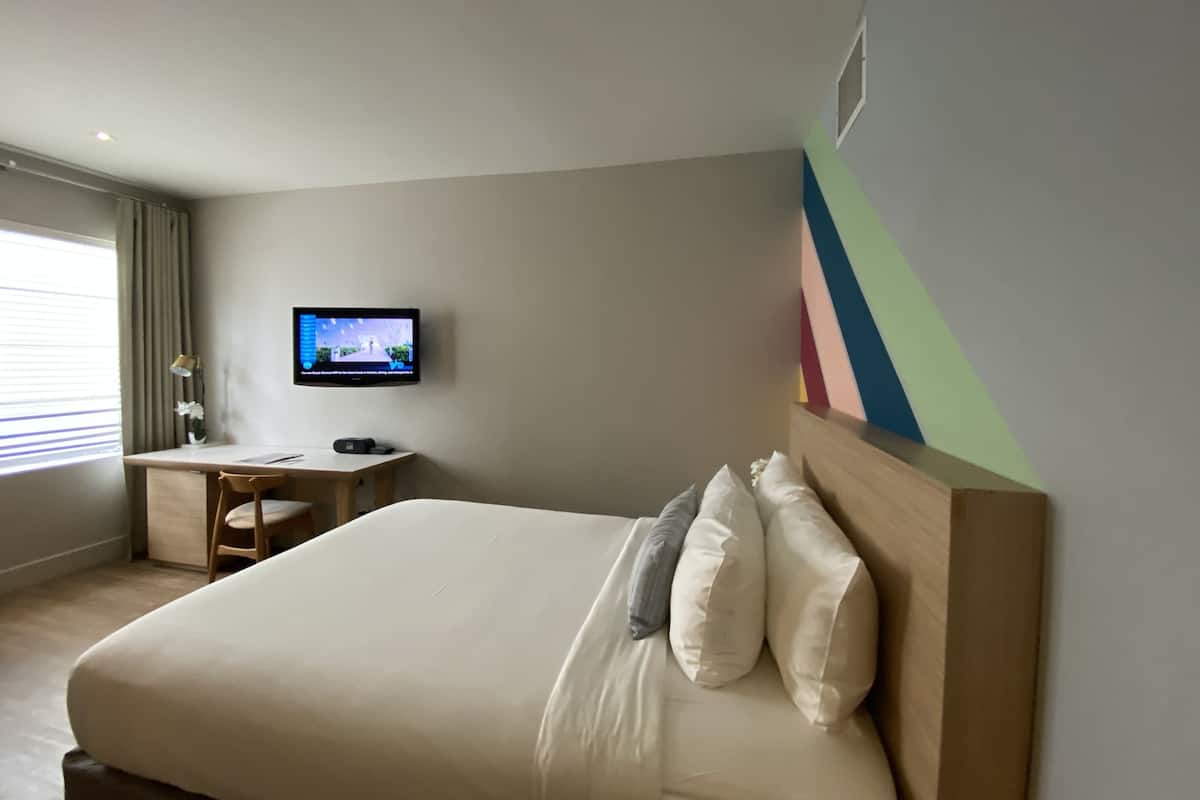 A hotel room with a bed, a desk, a chair, a TV, and a colorful wall.