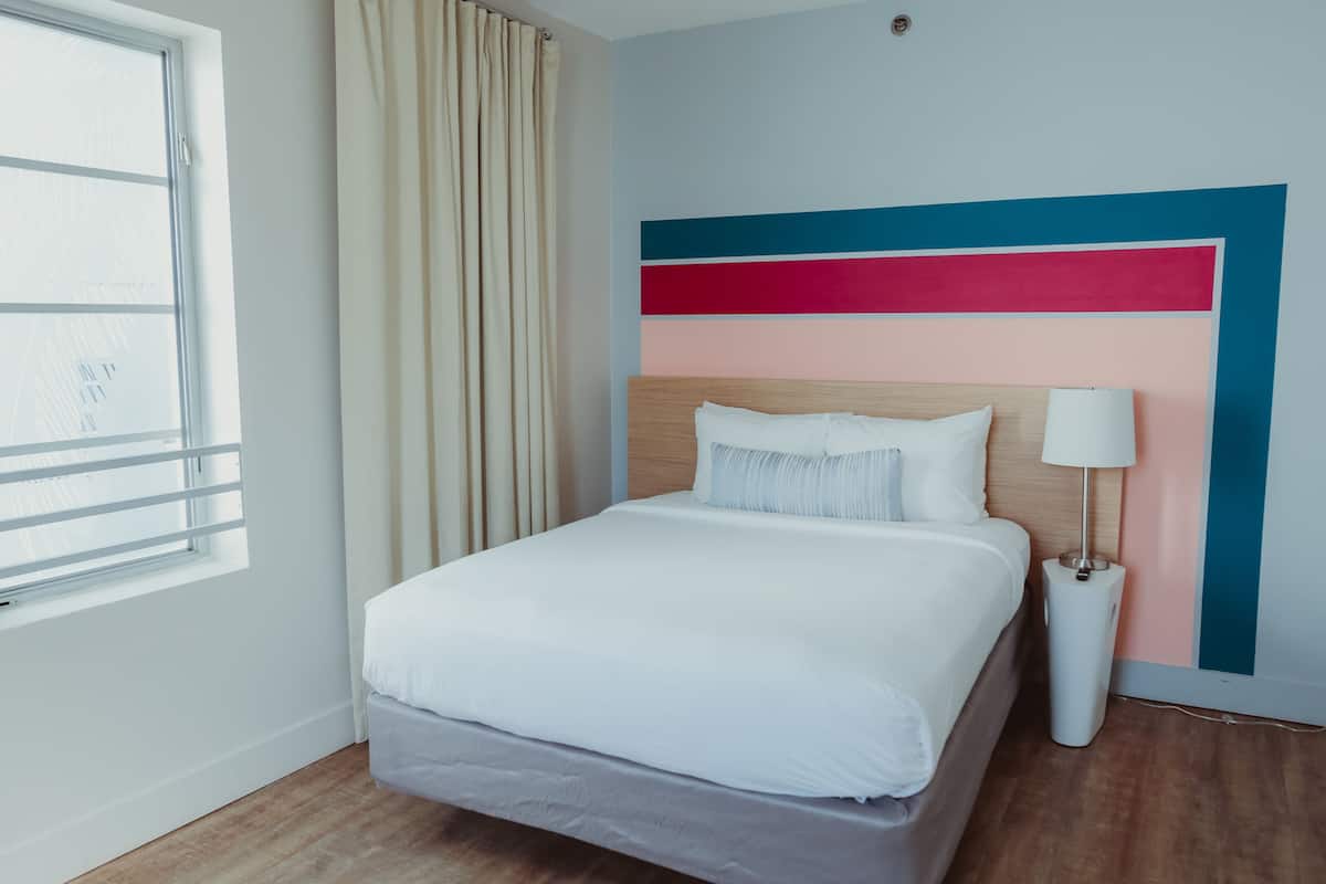 A neatly made bed with white linens, a bedside lamp, and a window with curtains.