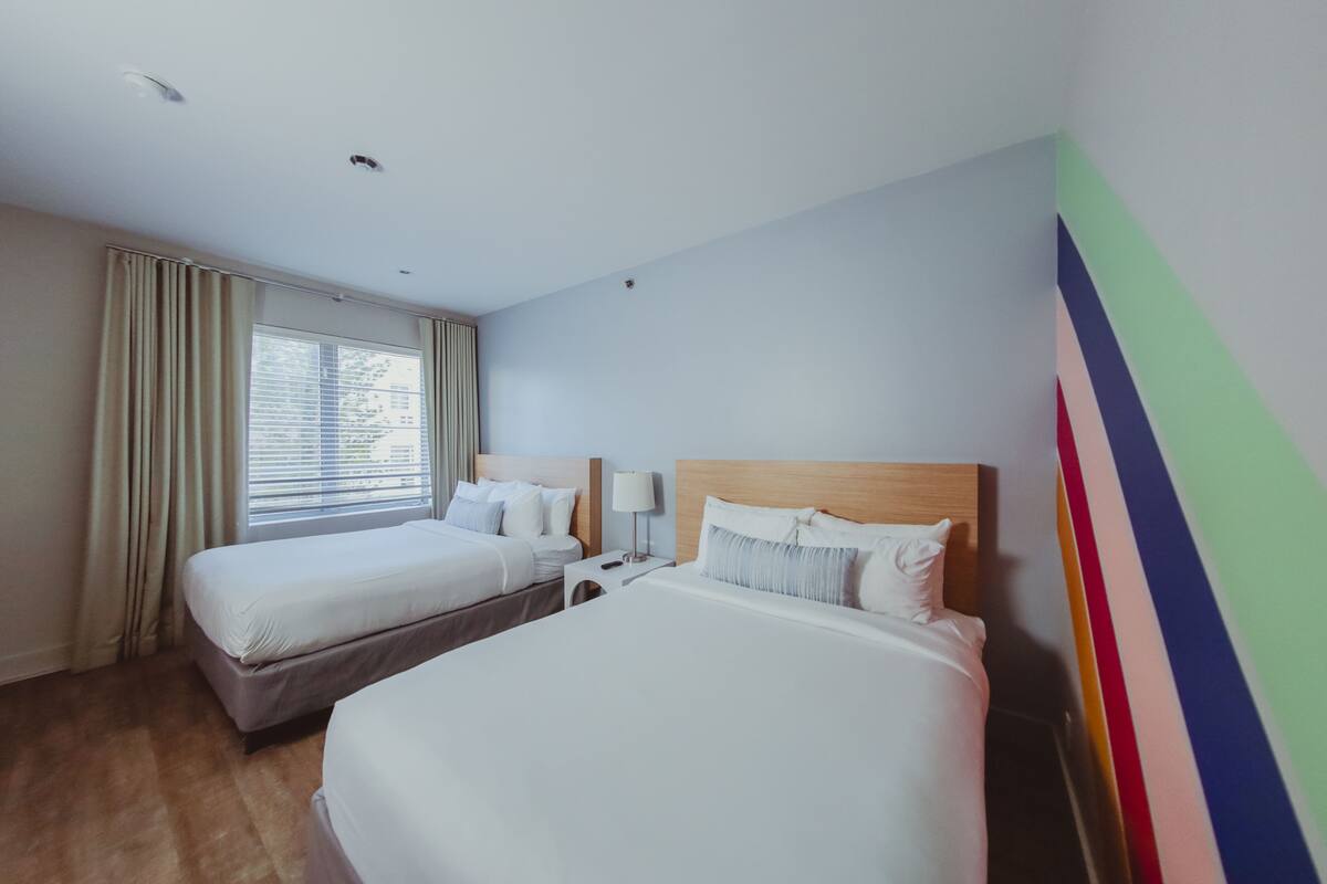 A hotel room with two beds, a window with curtains, a nightstand with a lamp, and a colorful wall decoration.