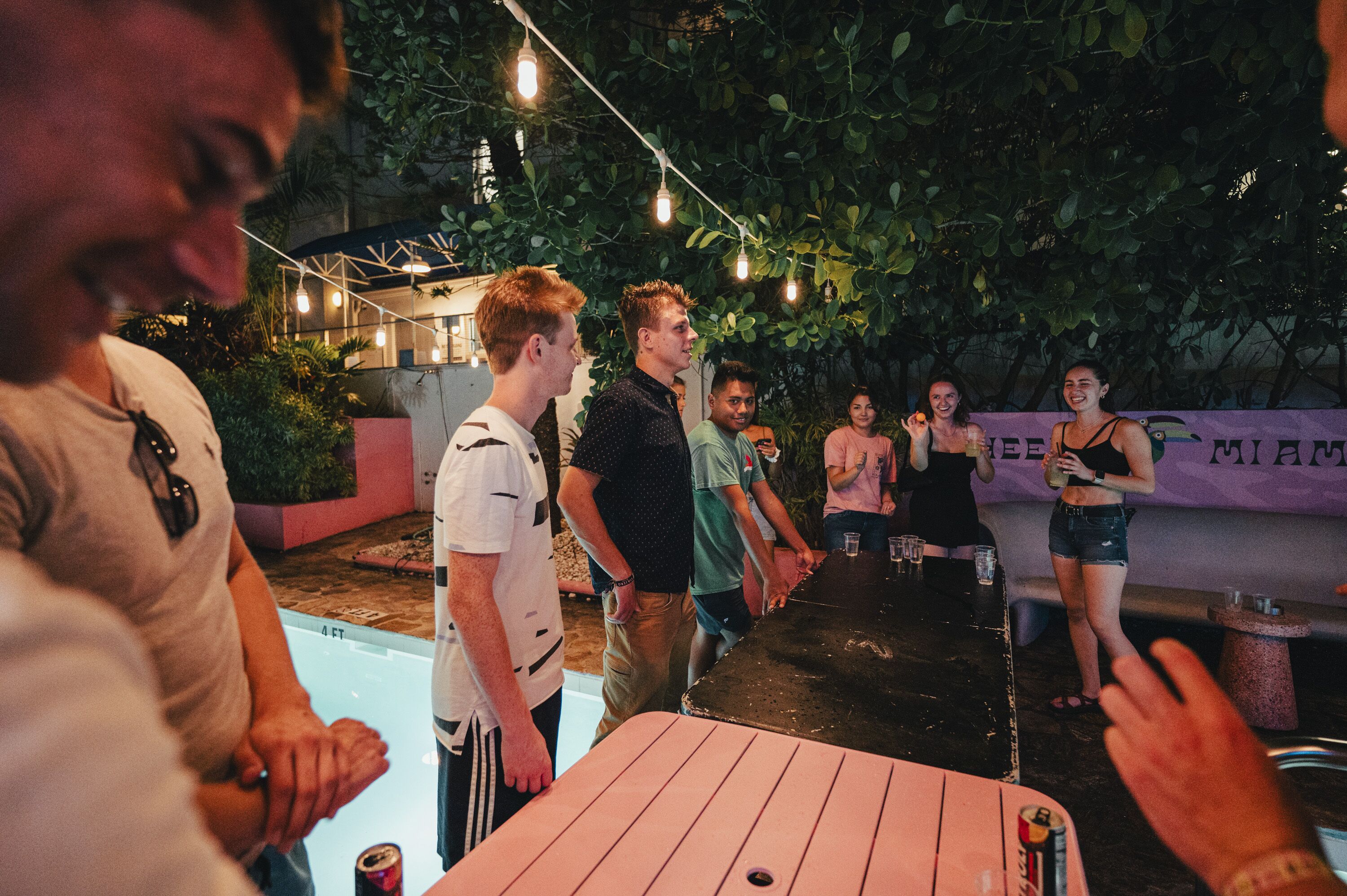 Un groupe de personnes se trouve autour d’une table de billard dans un espace extérieur, avec des guirlandes lumineuses suspendues au-dessus.