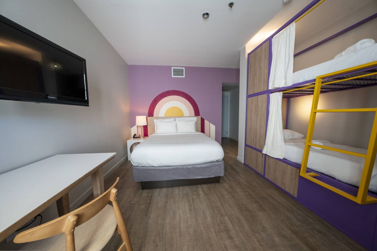 A hotel room with a bed, a desk, a chair, a TV, and bunk beds.