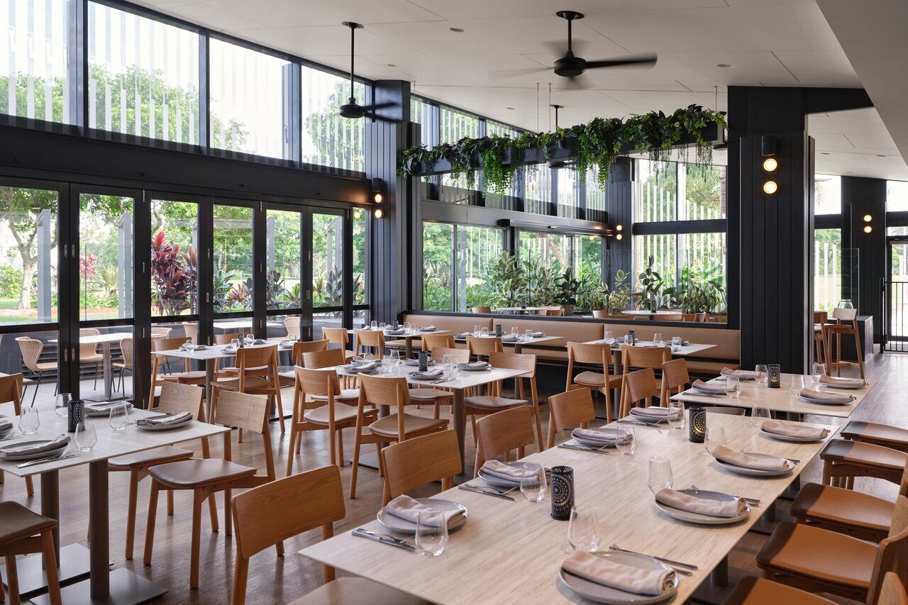A spacious restaurant with long wooden tables and chairs, large windows, and hanging plants.