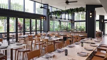 A spacious restaurant with long wooden tables and chairs, large windows, and hanging plants.