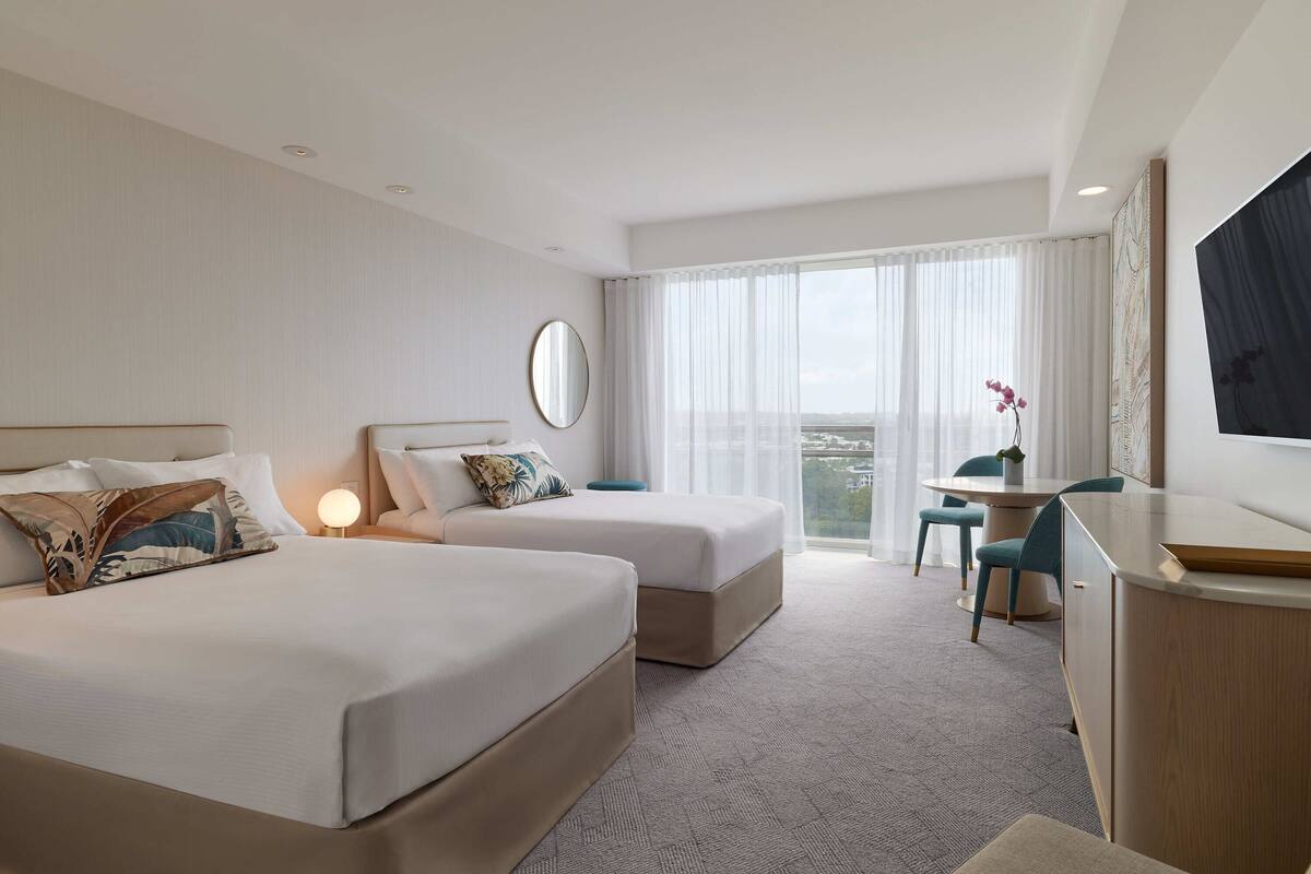A hotel room with two beds, a desk, a chair, a TV, and a large window with curtains.