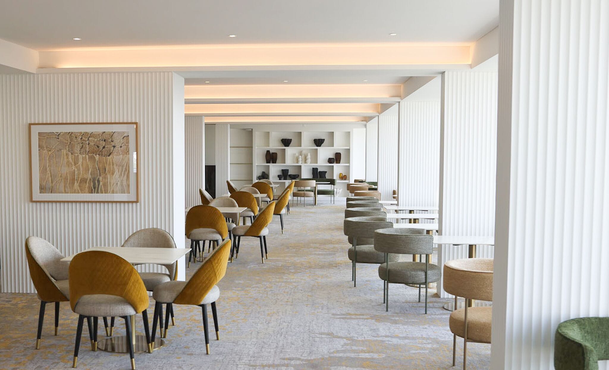 A modern hotel lobby with a long row of tables and chairs, a central art piece, and a patterned carpet.