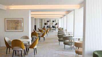 A modern hotel lobby with a long row of tables and chairs, a central art piece, and a patterned carpet.