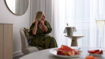 A woman in a green dress sitting on a sofa in a room with a round mirror, a small table with a bottle, and plates of fruit.