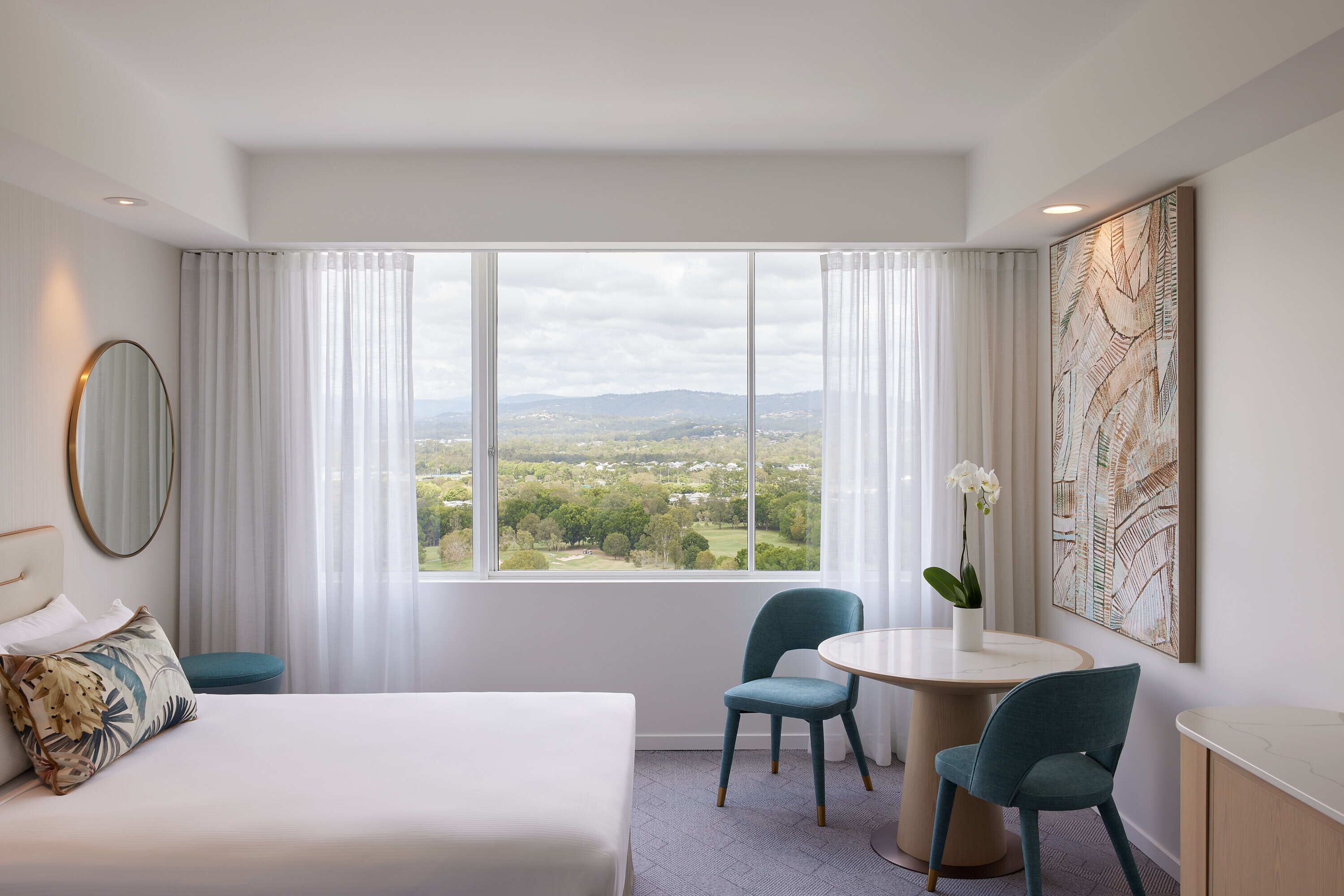 A hotel room with a large bed, a round table, two chairs, a view of the countryside, and a large abstract painting on the wall.