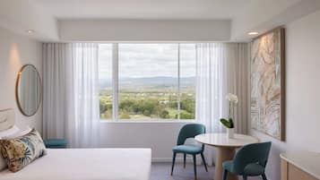 A hotel room with a large bed, a round table, two chairs, a view of the countryside, and a large abstract painting on the wall.