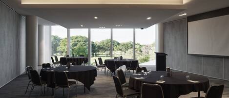 A conference room with round tables and chairs, large windows with a view of trees, and a projection screen on the wall.