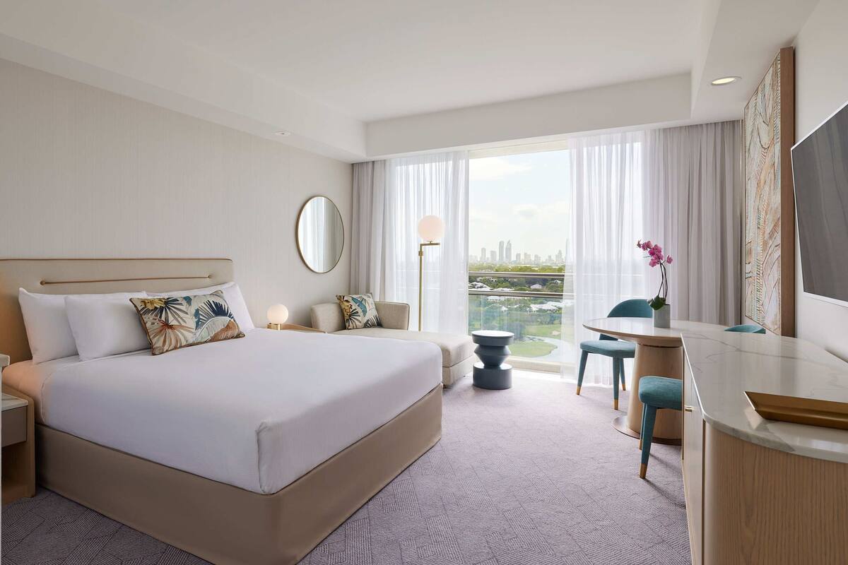 A hotel room with a large bed, a desk, a chair, a round table, and a view of the cityscape through the window.
