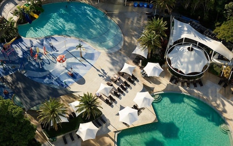An aerial view of a resort with multiple swimming pools, sun loungers, umbrellas, and palm trees.