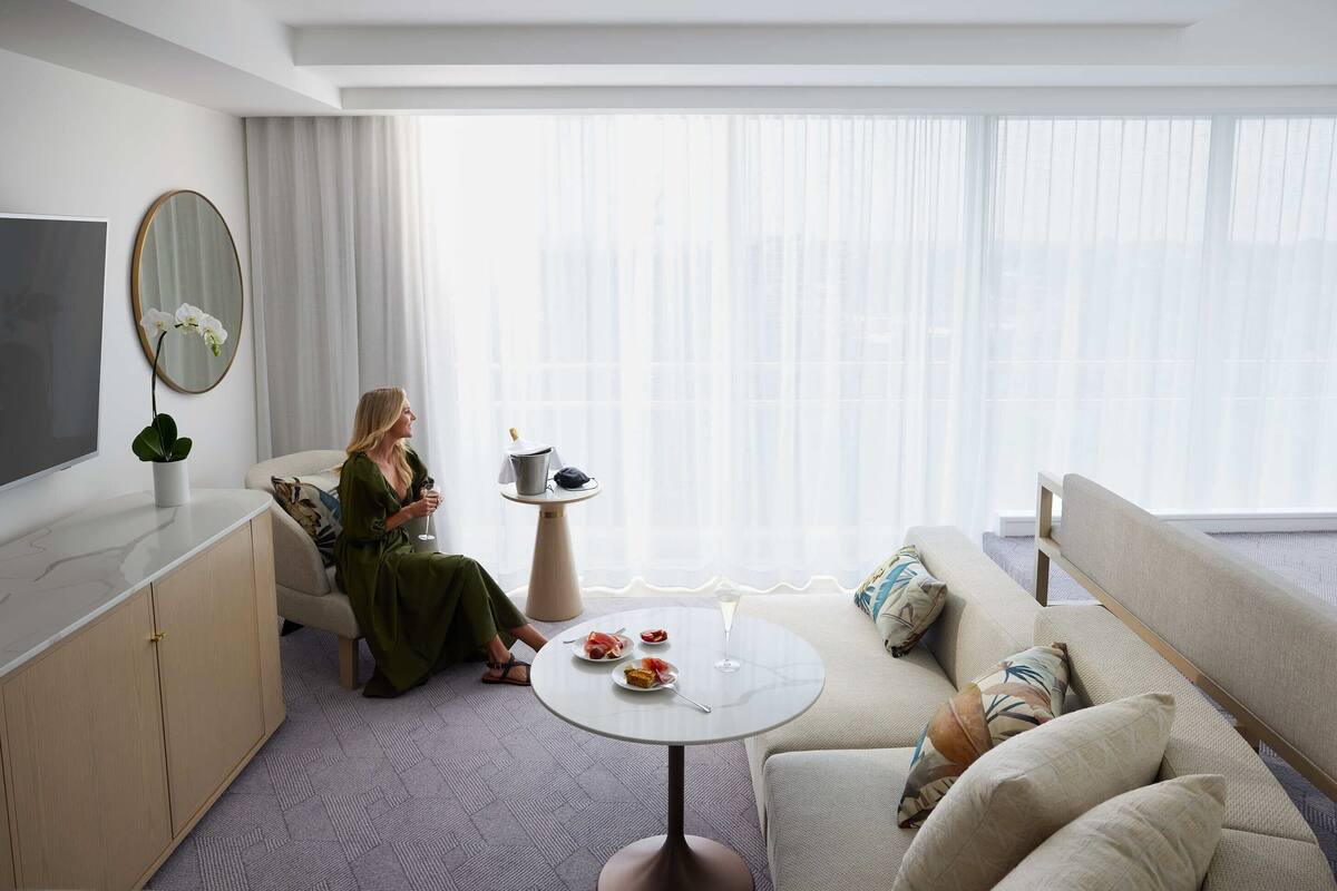 A modern hotel room with a sofa, a small round table with a drink, a TV, and a large window with sheer curtains.