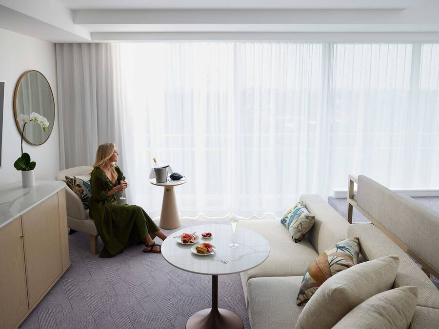 Una habitación de hotel moderna con un sofá, una mesita redonda con una bebida, un televisor y un ventanal con cortinas traslúcidas.