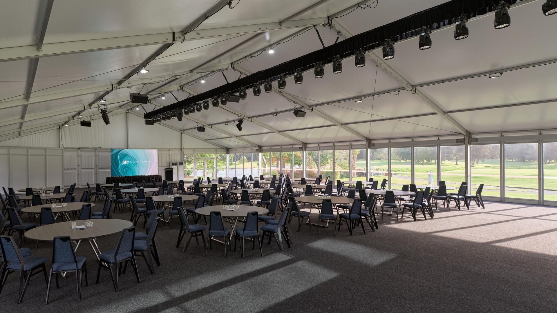 A large tented conference room with tables and chairs, a projection screen, and a view of a golf course.