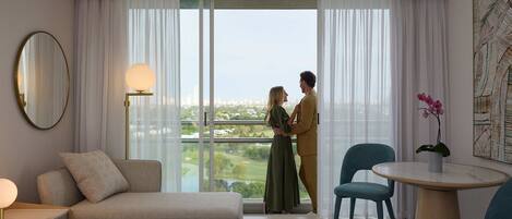 A hotel room with a large window, a bed, a sofa, a round table, and a chair.