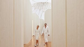 Un couloir au sol en bois et aux murs blancs, qui mène à un grand espace ouvert doté d’une structure de plafond aux formes géométriques.