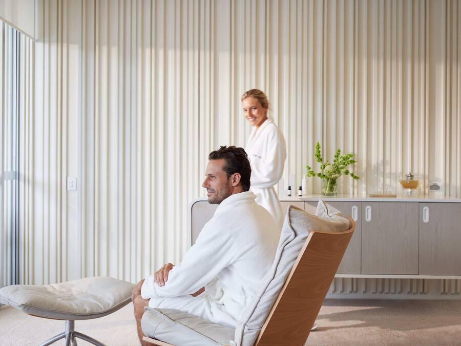 Ein moderner Spa-Bereich mit großem Fenster, einem gepolsterten Sessel, einer Holdekor-Kommode mit Pflegeprodukten und einer Person in einem Bademantel.