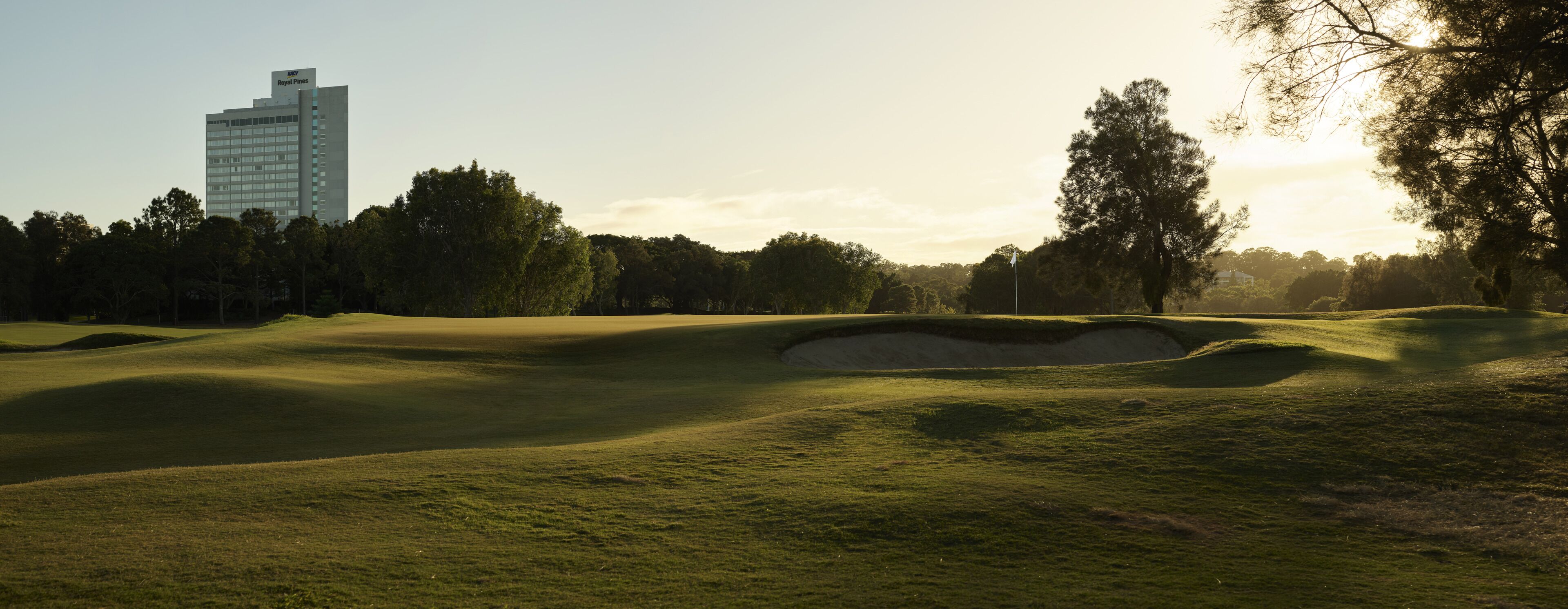 Un parcours de golf avec des bunkers et des arbres, et un bâtiment portant l’inscription « Royal Pines » en arrière-plan.