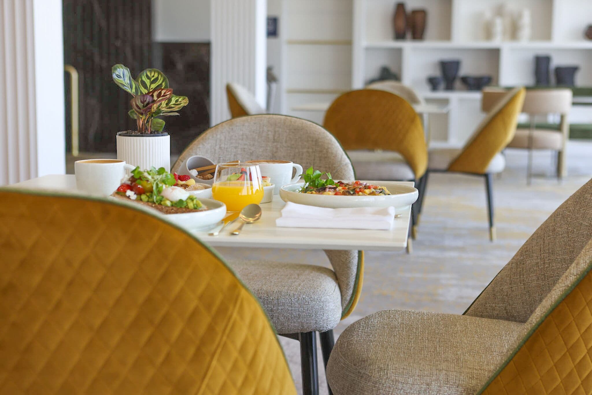 A dining area with a table set for breakfast, including plates of food, a glass of orange juice, and a coffee cup. There are chairs around the table, and a plant is visible in the background.