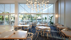 Un vaste espace de restauration intérieur, doté de grandes fenêtres, de tables et de chaises en bois, agrémenté d’un lustre moderne et offrant une vue sur la terrasse extérieure.
