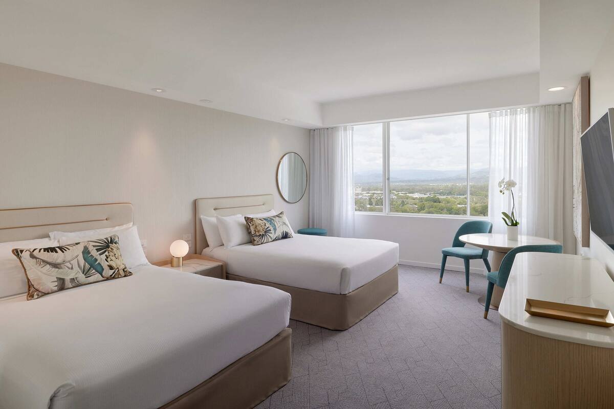 A hotel room with two beds, a desk, a chair, a large window with a view, and a round mirror on the wall.