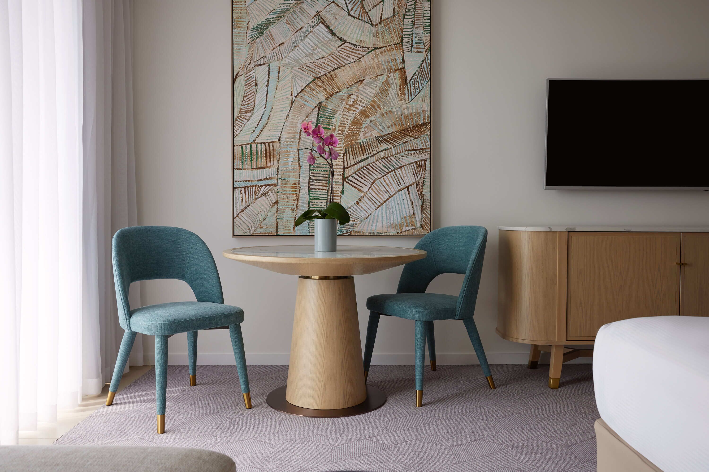 A hotel room with a round wooden table, two teal chairs, a potted orchid, a large abstract painting, a TV, and a wooden cabinet.