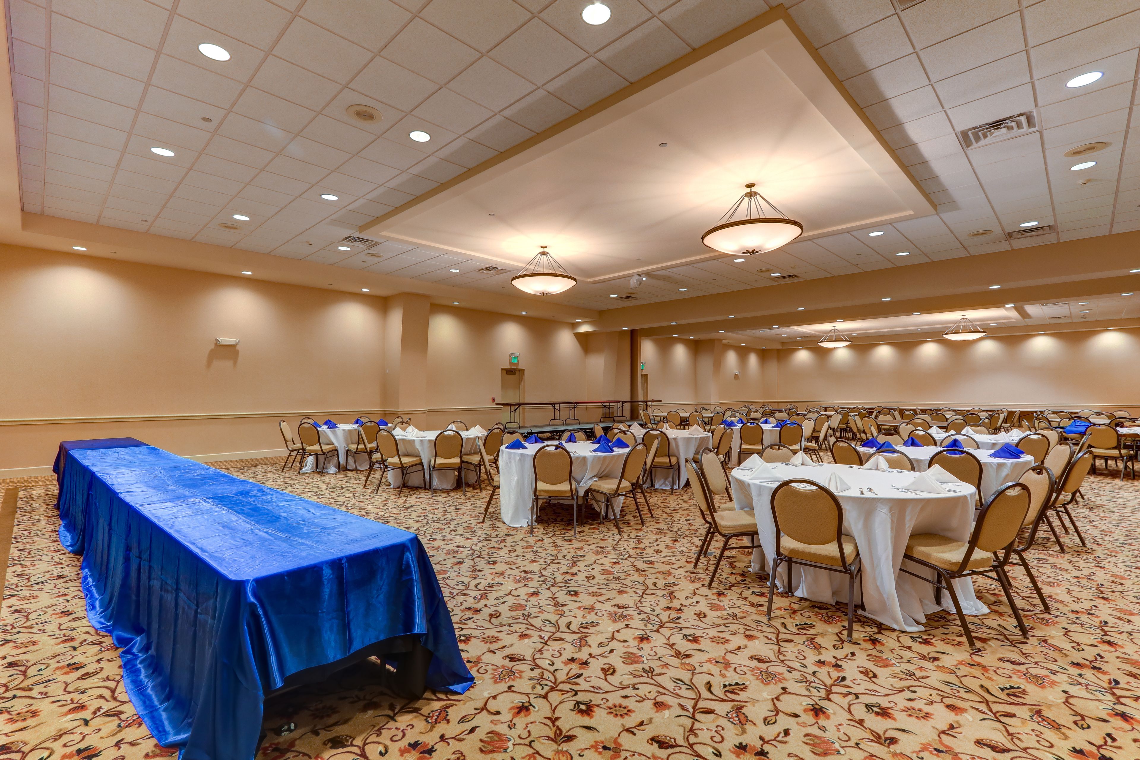 A banquet hall with rows of round tables and chairs, a long table with a blue tablecloth, and a carpeted floor.