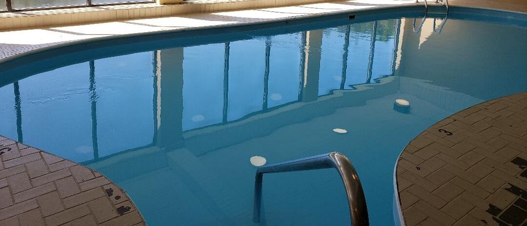 An indoor swimming pool with clear water, a metal ladder, and tiled flooring.