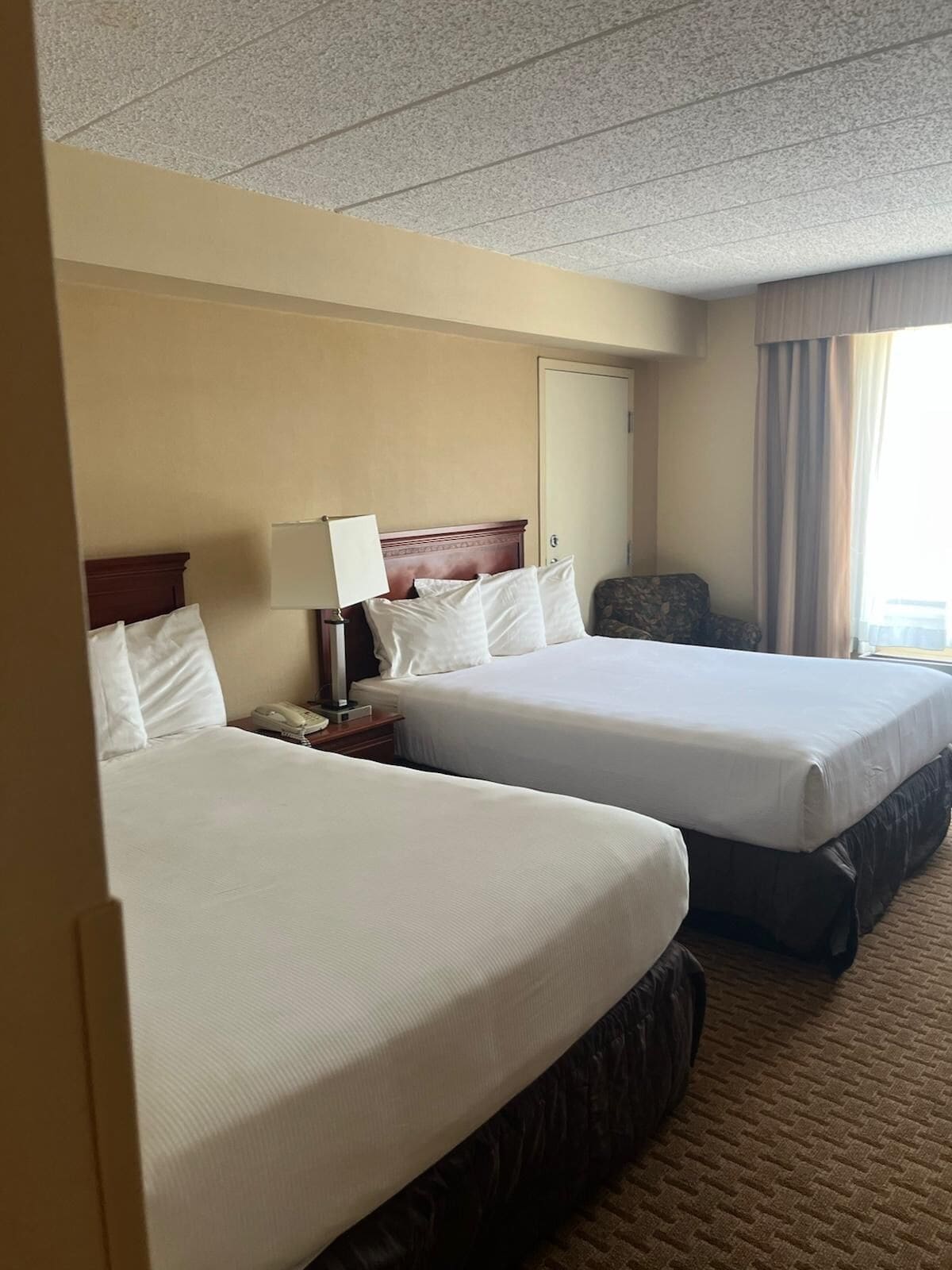 A hotel room with two beds, a nightstand with a lamp, a chair, and a window with curtains.