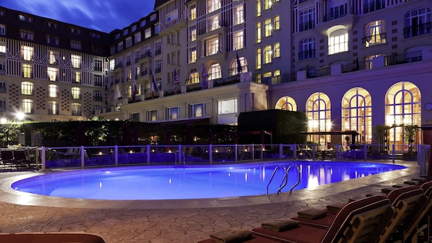 A nighttime swimming pool area with lounge chairs and a building with multiple balconies in the background. - Hôtel Barrière Le Royal Deauville (Deauville)