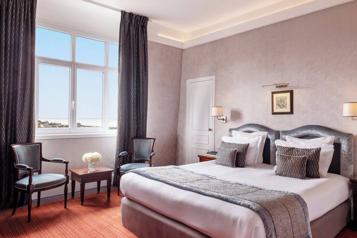 Et hotelværelse med en stor seng, to stole, et natbord og et vindue med gardiner.