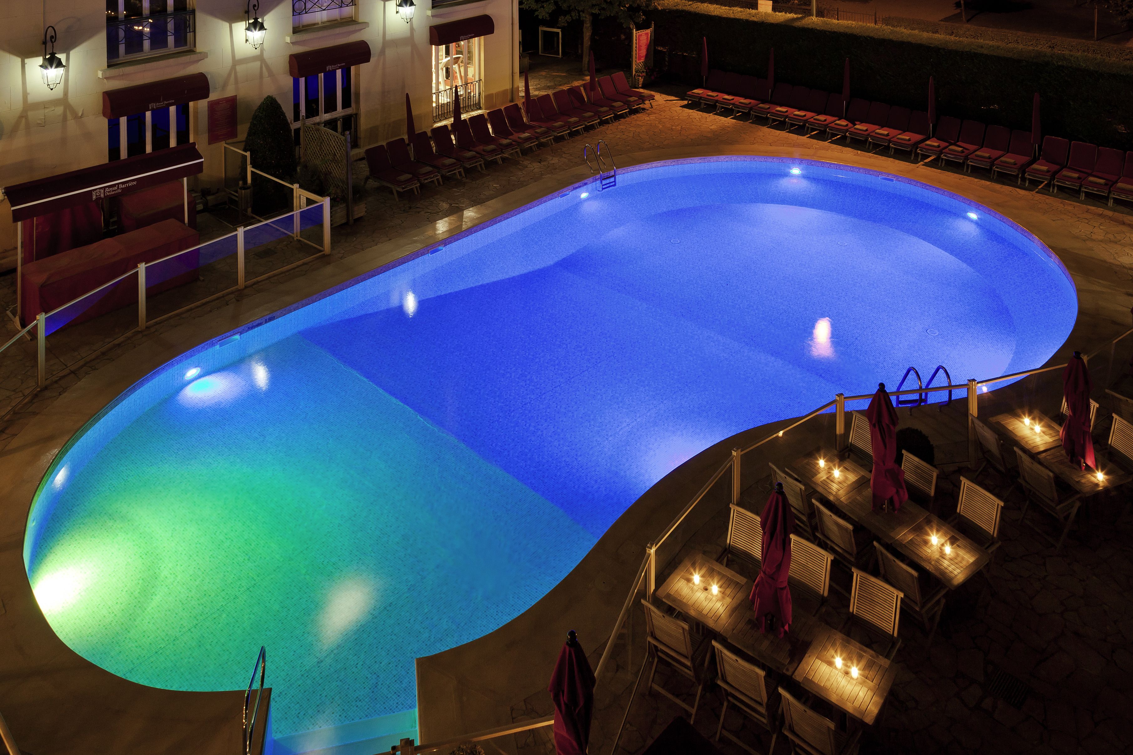 A large indoor swimming pool with a curved edge, surrounded by red chairs and tables with umbrellas.