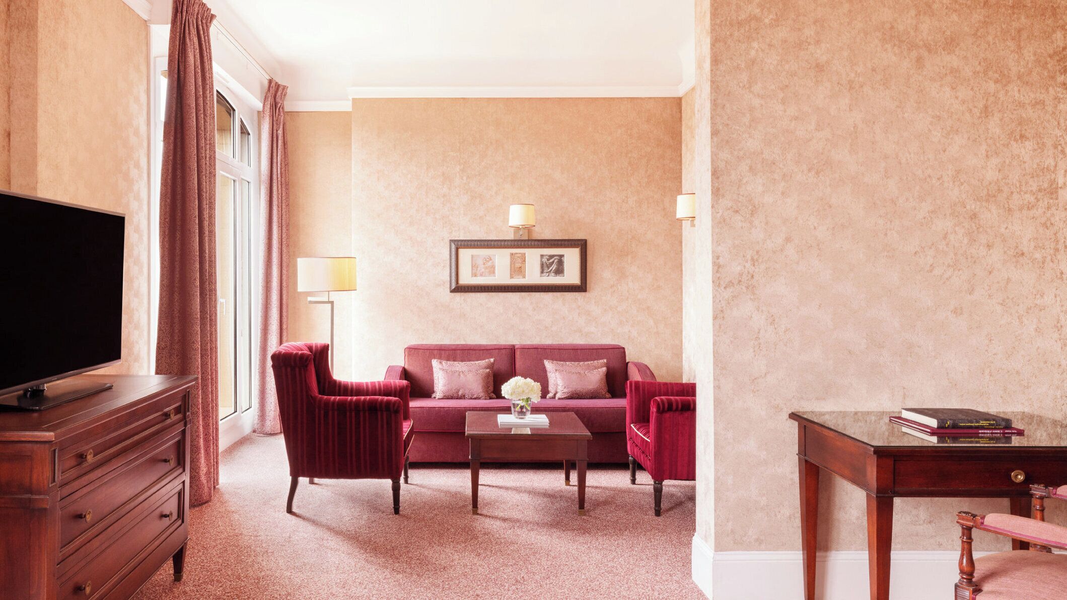 A hotel room with a large flat-screen TV, a red sofa, a wooden coffee table, and a small side table with books.