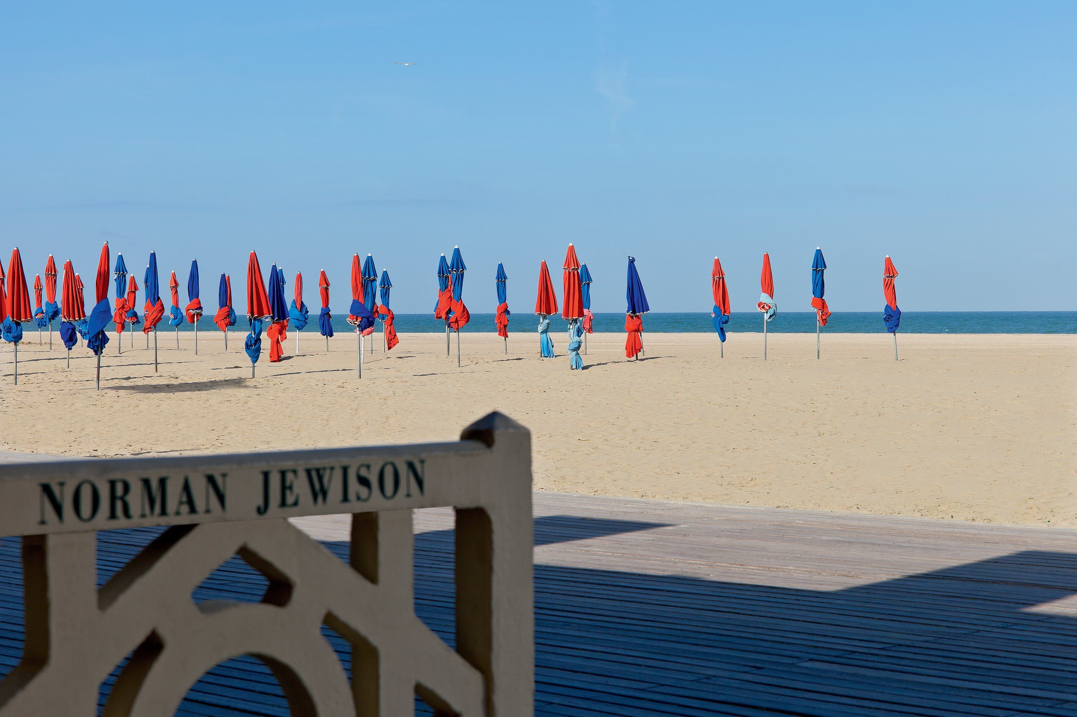 A beach with rows of red and blue umbrellas, a wooden boardwalk, and a sign that reads "Norman Jewison".
