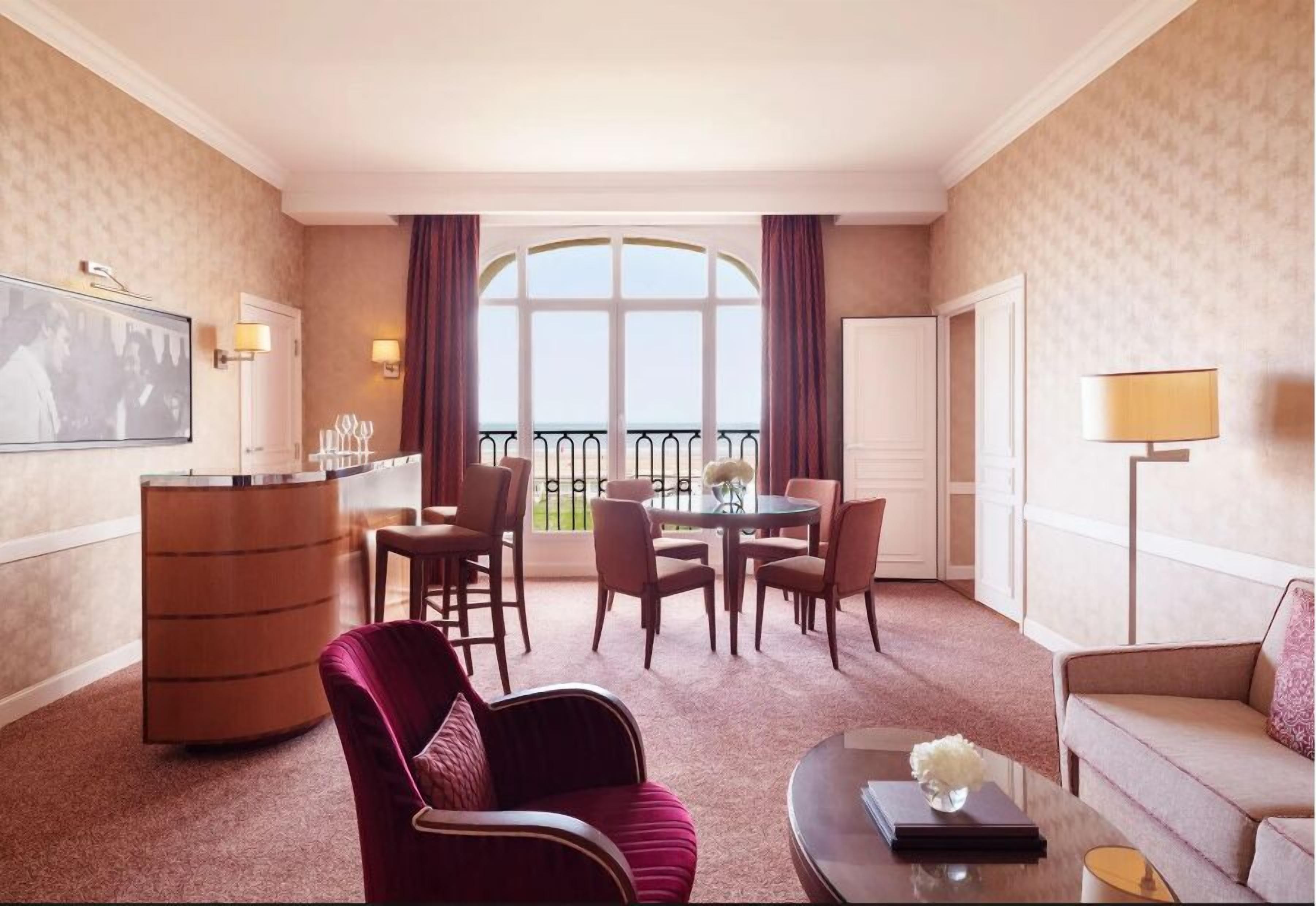 A hotel room with a dining area, a large window with a view, a red armchair, a sofa, and a small table with a vase.
