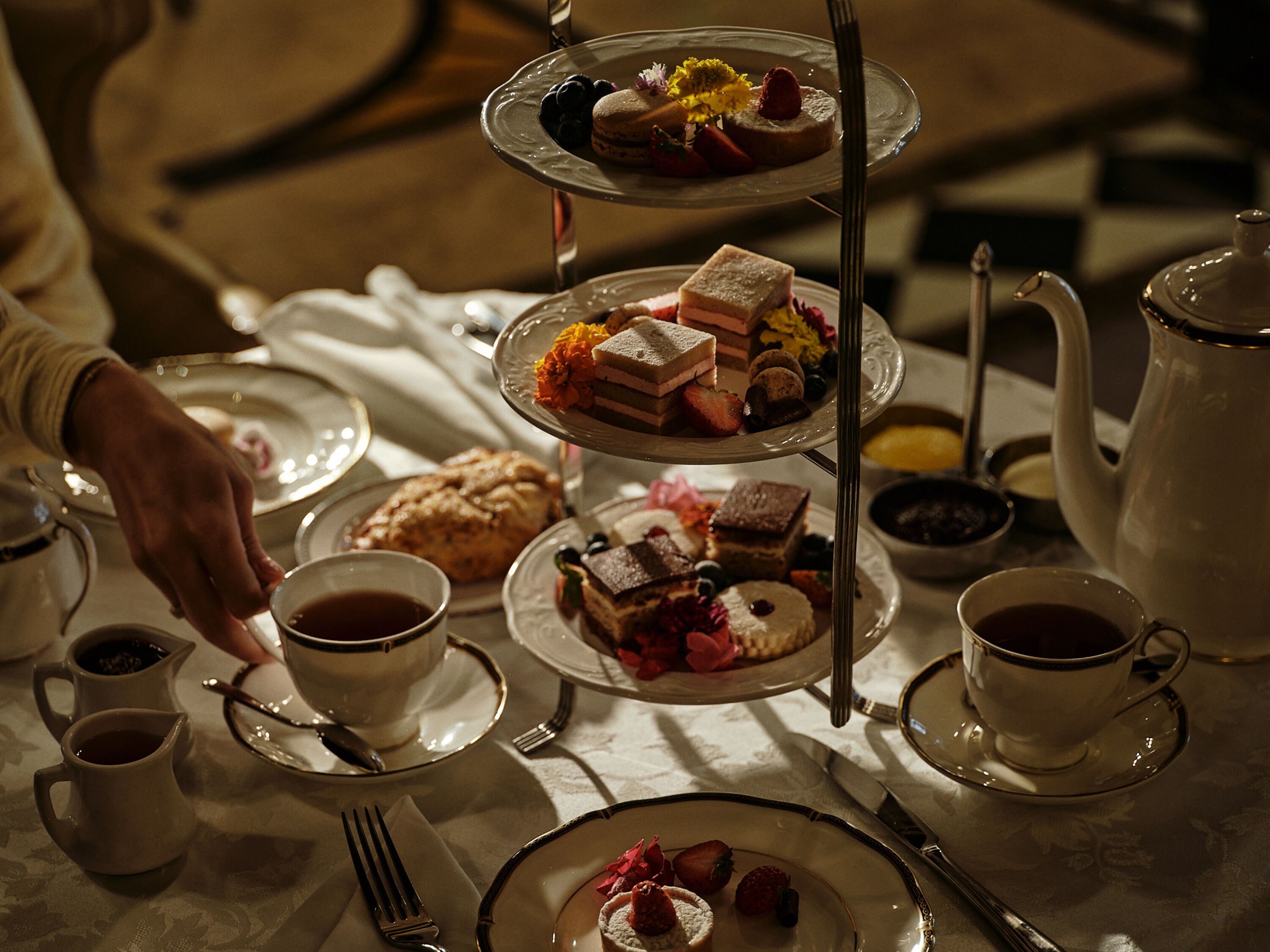 A three-tiered dessert stand with various pastries and fruits, accompanied by cups of tea and a teapot on a table.