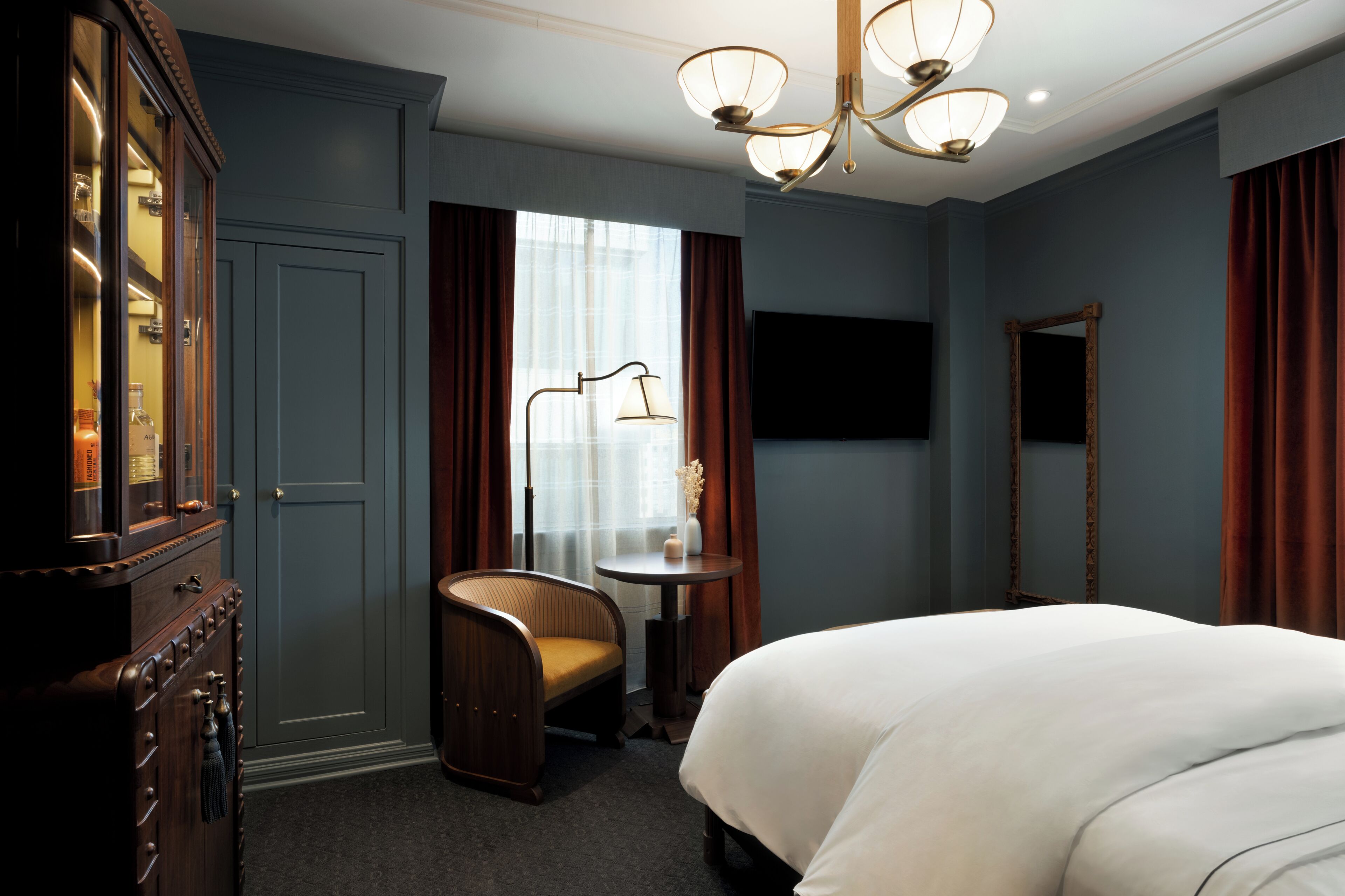 Une chambre d’hôtel avec un lit, une commode en bois, une chaise, une petite table, un téléviseur et un lustre.
