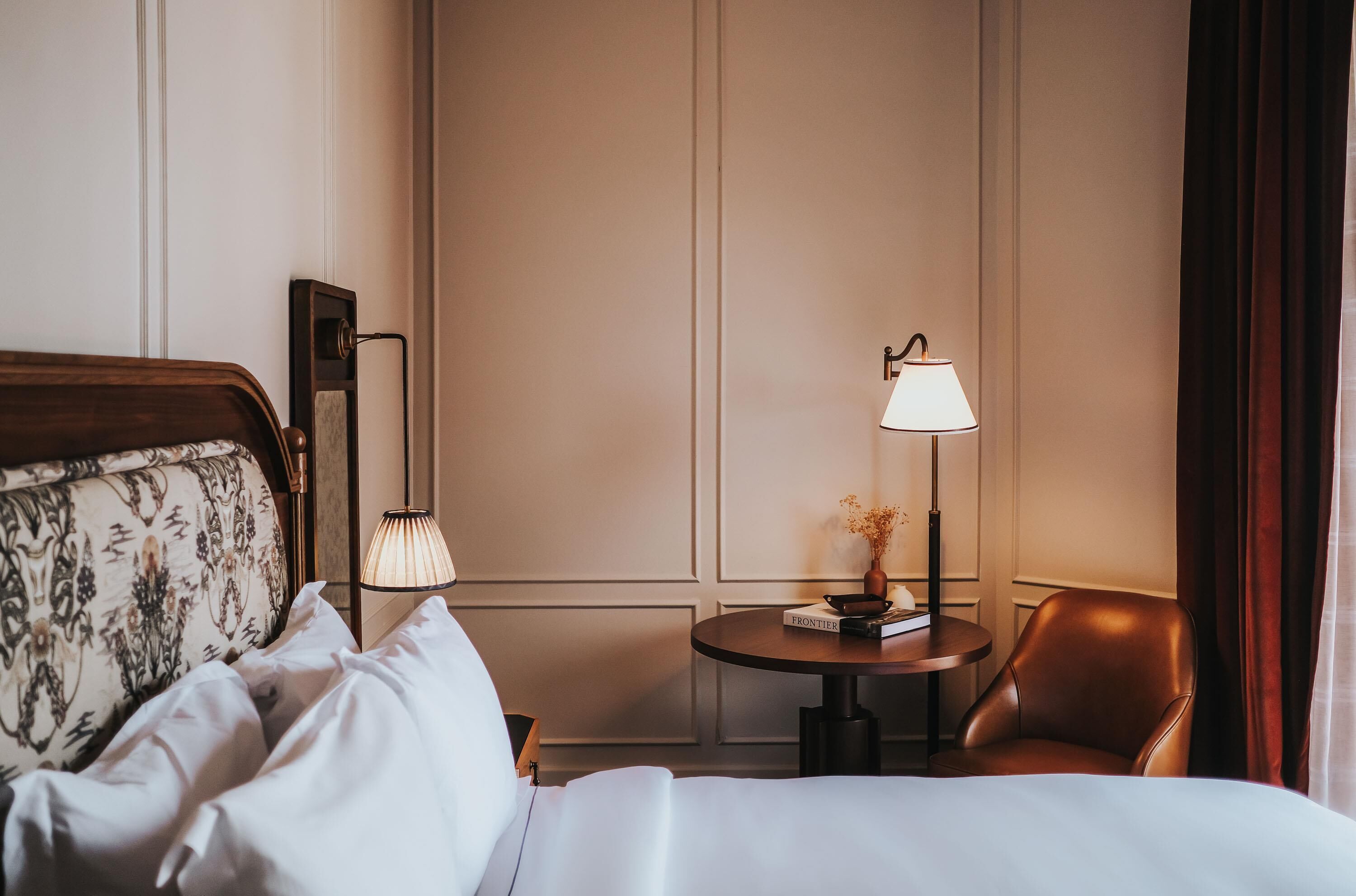 A hotel room with a bed, a chair, a table with a lamp, and a bookshelf.