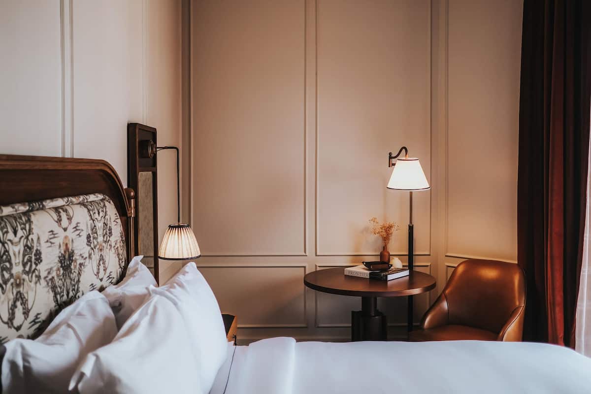 A hotel room with a bed, a chair, a table with a lamp, and a bookshelf.