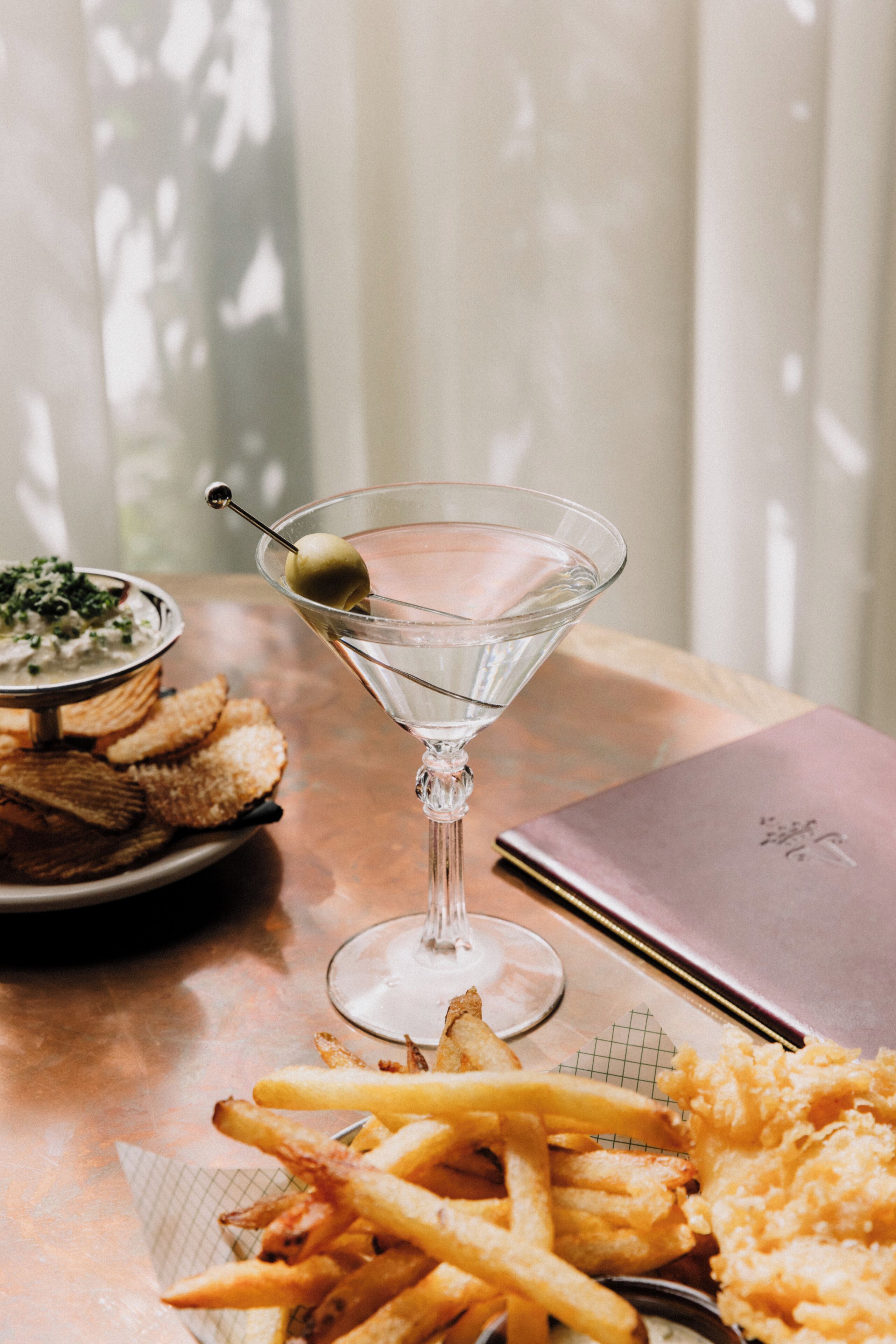 A martini glass with an olive, a plate of French fries, and a bowl of dip on a table.