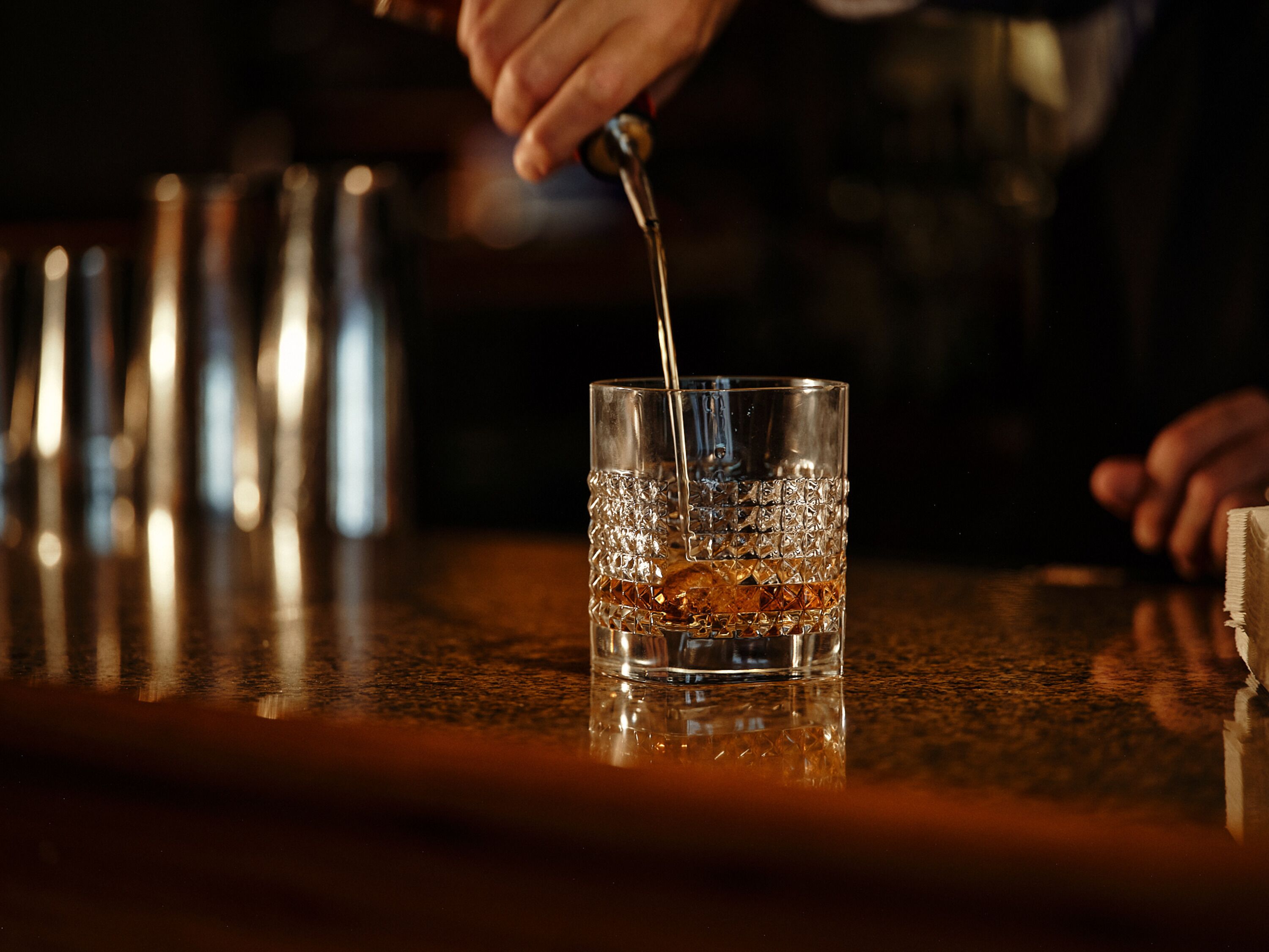 A bar setting with a glass being filled with a dark liquid, ice cubes, and a bar tool.