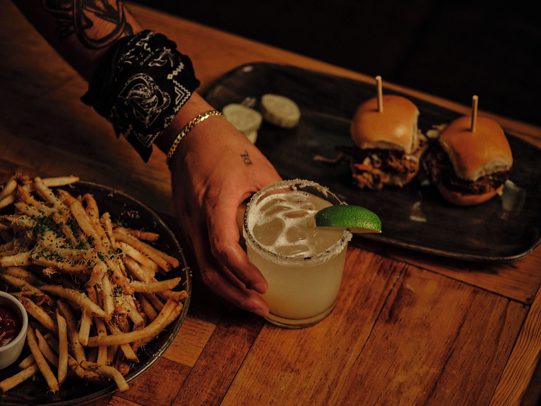 A hand holding a margarita with a lime garnish, a plate of seasoned fries, and a burger on a tray.